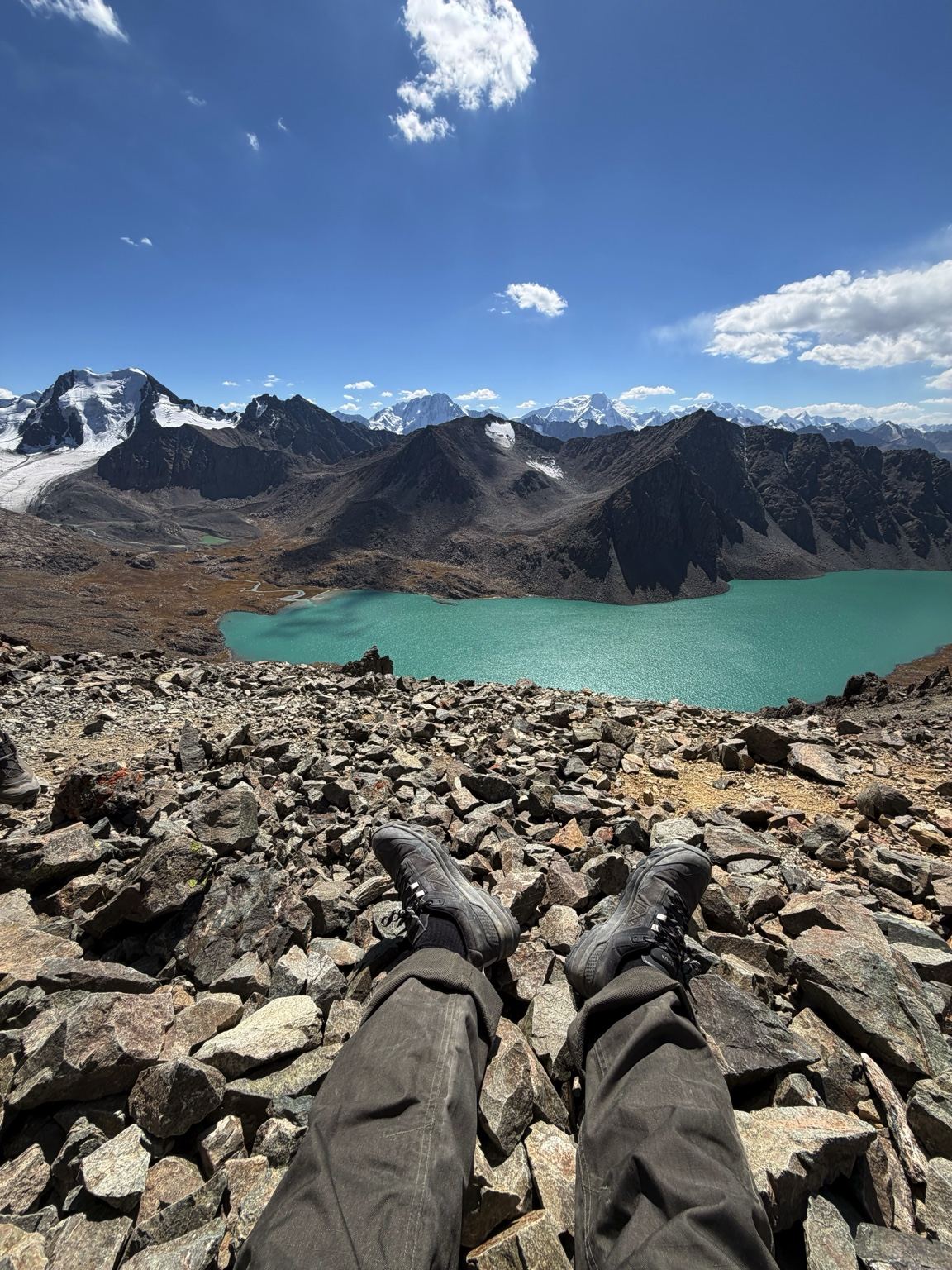Wandelschoenen op een rotsige helling met uitzicht op een turquoise bergmeer en besneeuwde toppen