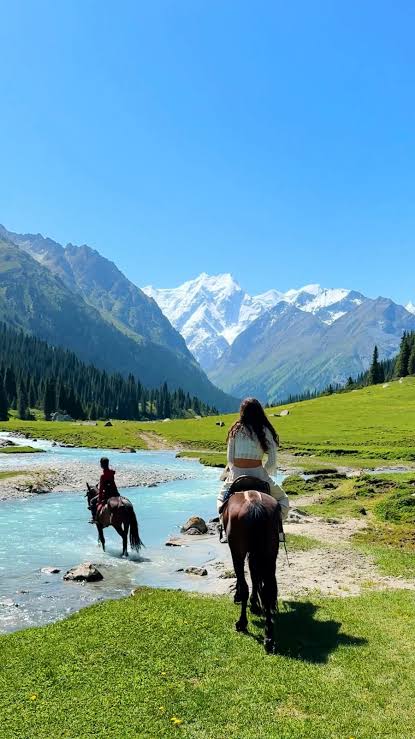 Twee reizigers te paard door een turquoise bergbeek, met groene weiden en besneeuwde toppen op de achtergrond